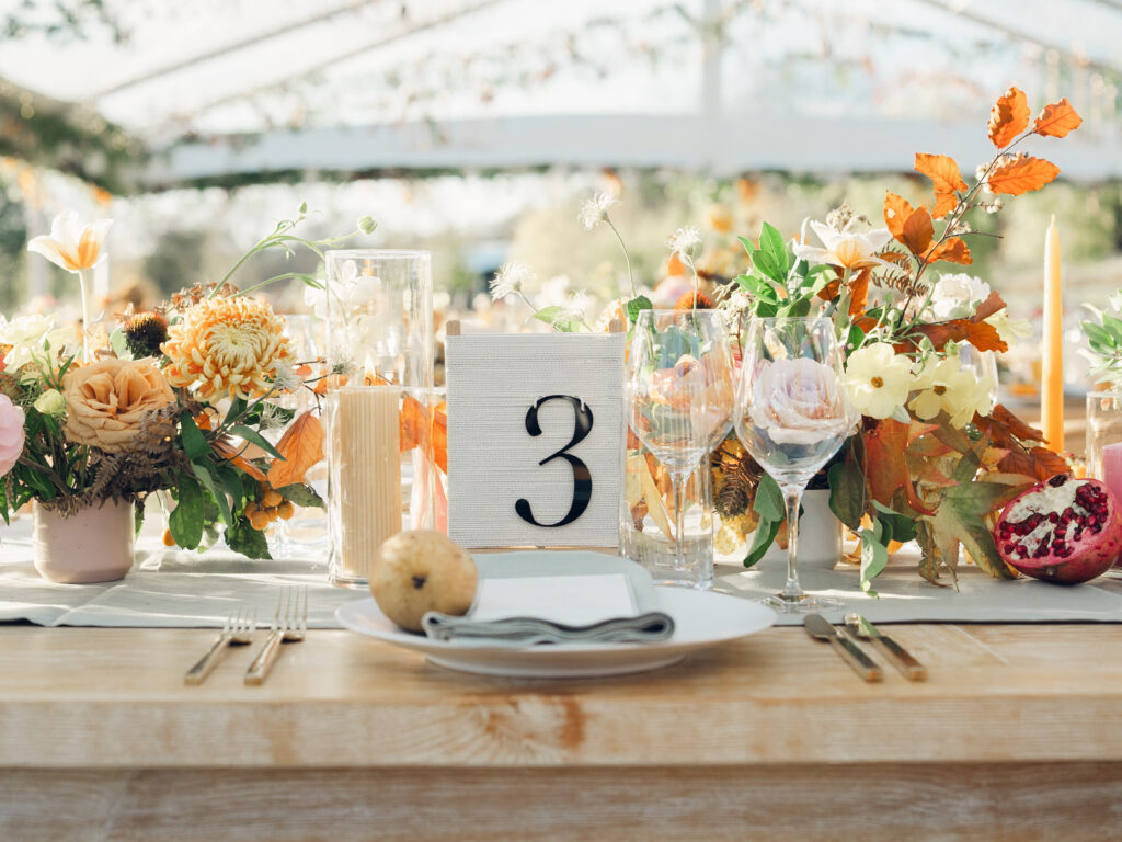 Full tablescapes of fall flowers create lush warm fall setting for wedding reception in Tennessee countryside. Wedding colors in blush, tangerine, mustard yellow, caramel, and honey. Wedding fall florals consisting of roses, clematis, ranunculus, brown tulips, and fall foliage. A collection of low centerpieces and accent arrangements create lush floral wedding reception tables with styled fruit place settings. Unique and colorful candles for reception tables in pink, orange, yellow, and gold. Destination floral design by Rosemary & Finch Floral Design in Nashville, TN.