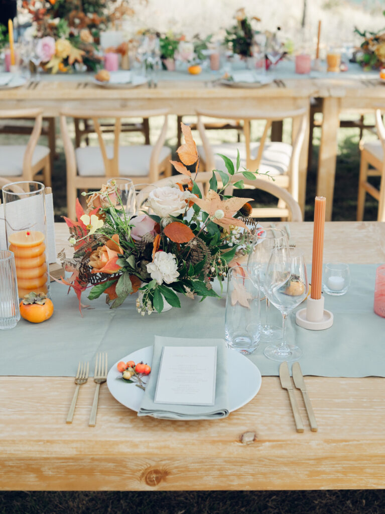 Full tablescapes of fall flowers create lush warm fall setting for wedding reception in Tennessee countryside. Wedding colors in blush, tangerine, mustard yellow, caramel, and honey. Wedding fall florals consisting of roses, clematis, ranunculus, brown tulips, and fall foliage. A collection of low centerpieces and accent arrangements create lush floral wedding reception tables with styled fruit place settings. Unique and colorful candles for reception tables in pink, orange, yellow, and gold. Destination floral design by Rosemary & Finch Floral Design in Nashville, TN.