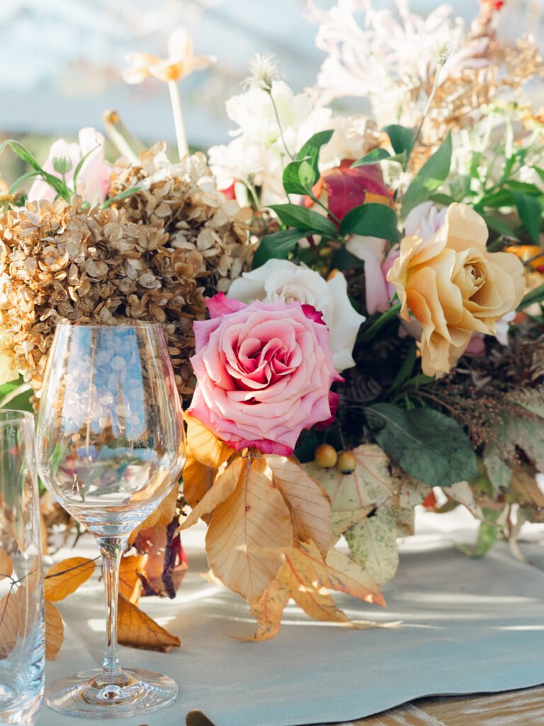 Full tablescapes of fall flowers create lush warm fall setting for wedding reception in Tennessee countryside. Wedding colors in blush, tangerine, mustard yellow, caramel, and honey. Wedding fall florals consisting of roses, clematis, ranunculus, brown tulips, and fall foliage. A collection of low centerpieces and accent arrangements create lush floral wedding reception tables with styled fruit place settings. Unique and colorful candles for reception tables in pink, orange, yellow, and gold. Destination floral design by Rosemary & Finch Floral Design in Nashville, TN.