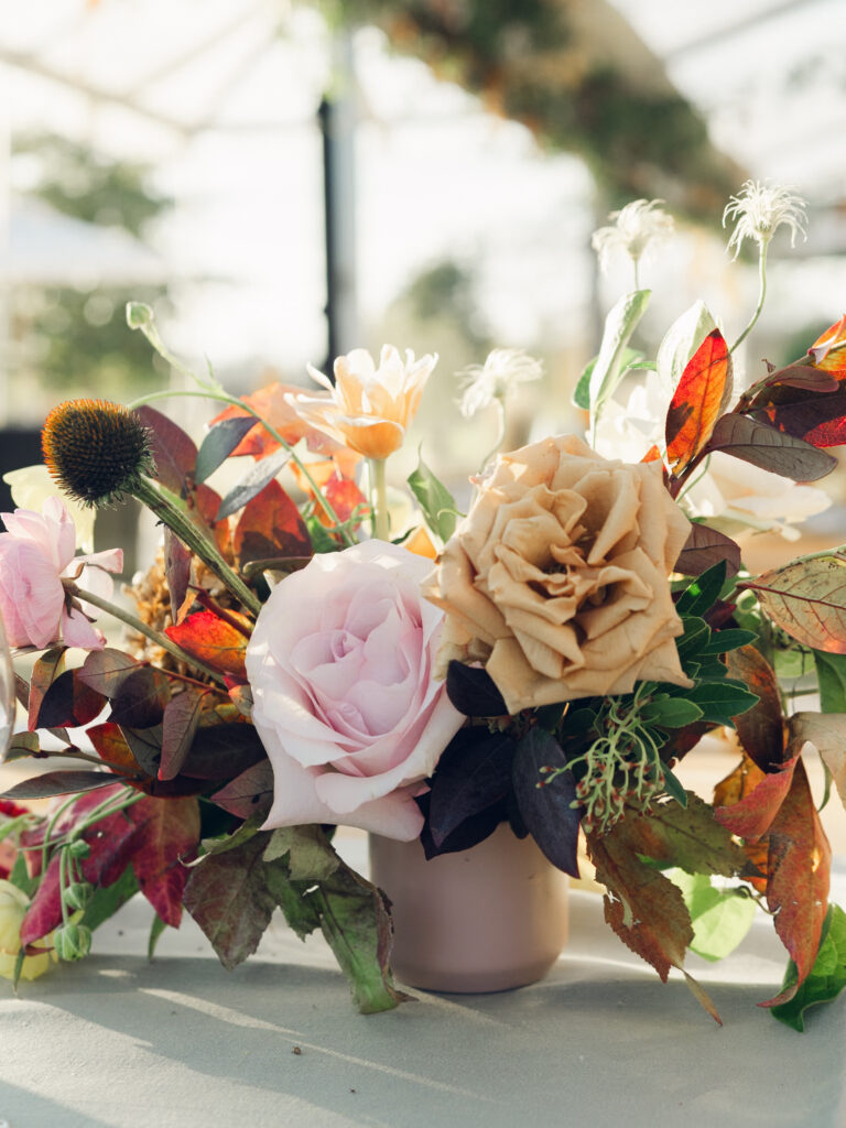 Full tablescapes of fall flowers create lush warm fall setting for wedding reception in Tennessee countryside. Wedding colors in blush, tangerine, mustard yellow, caramel, and honey. Wedding fall florals consisting of roses, clematis, ranunculus, brown tulips, and fall foliage. A collection of low centerpieces and accent arrangements create lush floral wedding reception tables with styled fruit place settings. Unique and colorful candles for reception tables in pink, orange, yellow, and gold. Destination floral design by Rosemary & Finch Floral Design in Nashville, TN.