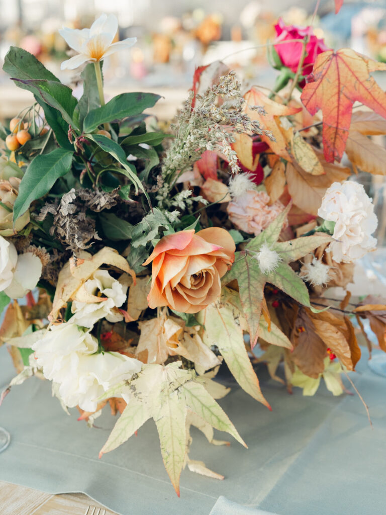 Full tablescapes of fall flowers create lush warm fall setting for wedding reception in Tennessee countryside. Wedding colors in blush, tangerine, mustard yellow, caramel, and honey. Wedding fall florals consisting of roses, clematis, ranunculus, brown tulips, and fall foliage. A collection of low centerpieces and accent arrangements create lush floral wedding reception tables with styled fruit place settings. Unique and colorful candles for reception tables in pink, orange, yellow, and gold. Destination floral design by Rosemary & Finch Floral Design in Nashville, TN.