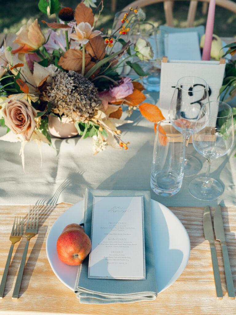 Full tablescapes of fall flowers create lush warm fall setting for wedding reception in Tennessee countryside. Wedding colors in blush, tangerine, mustard yellow, caramel, and honey. Wedding fall florals consisting of roses, clematis, ranunculus, brown tulips, and fall foliage. A collection of low centerpieces and accent arrangements create lush floral wedding reception tables with styled fruit place settings. Unique and colorful candles for reception tables in pink, orange, yellow, and gold. Destination floral design by Rosemary & Finch Floral Design in Nashville, TN.