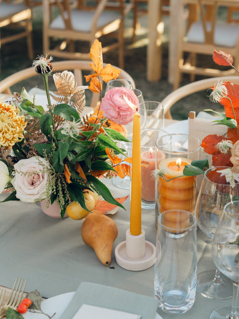 Full tablescapes of fall flowers create lush warm fall setting for wedding reception in Tennessee countryside. Wedding colors in blush, tangerine, mustard yellow, caramel, and honey. Wedding fall florals consisting of roses, clematis, ranunculus, brown tulips, and fall foliage. A collection of low centerpieces and accent arrangements create lush floral wedding reception tables with styled fruit place settings. Unique and colorful candles for reception tables in pink, orange, yellow, and gold. Destination floral design by Rosemary & Finch Floral Design in Nashville, TN.