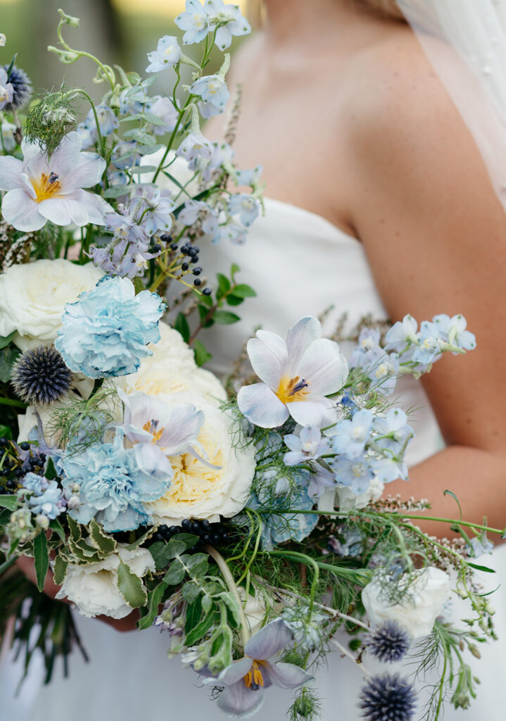 Whimsical bridal bouquet for summer wedding in blue and white. Garden-inspired bridal bouquet consisting or roses, blue tulips, nandina, carnations, ranunculus, and echinops. Medium-sized bridal bouquet for summer wedding. Design by Rosemary & Finch Floral Design in Nashville, TN. 