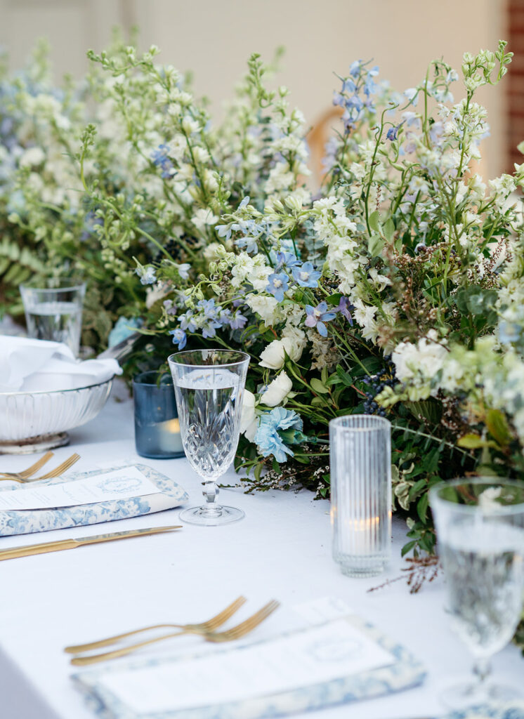 Long floral meadows line the head tables of this garden-inspired wedding at Cheekwood. Lush floral centerpieces in blue and white for summer wedding reception. White wedding florals with hint of blue consisting of white and blue delphinium. Clear and dusty blue candles add warm lighting for outdoor wedding reception. Floral table runner for wedding reception head tables. Design by Rosemary & Finch Floral Design in Nashville, TN.