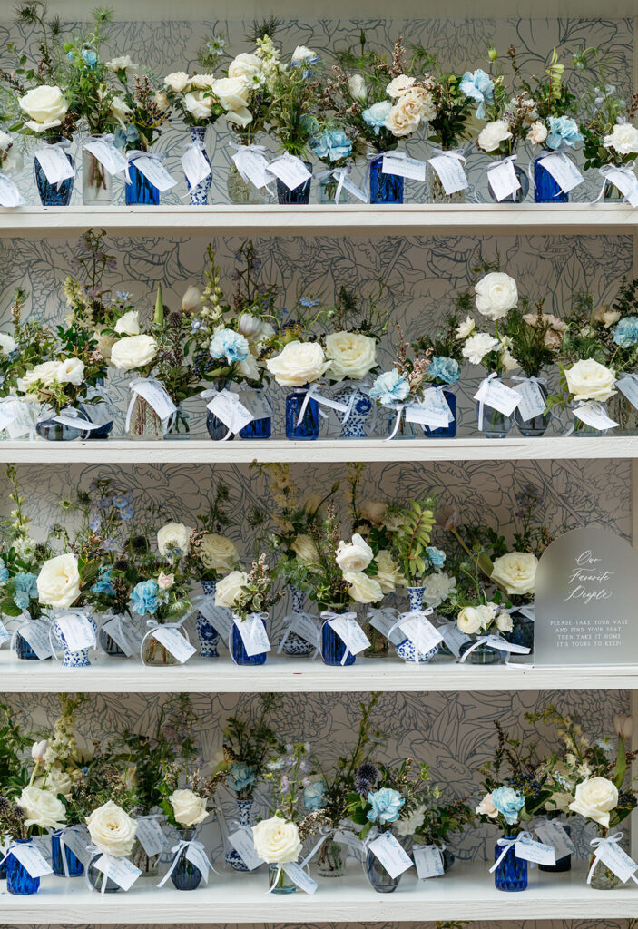 Bud Vase escort display in colors blue and white for summer wedding reception. Antique blue and white vases decorate summer wedding escort display with roses, ranunculus, carnations, and greenery. Escort display with floral party favors for guests. Italian inspired wedding reception with vintage blue and white floral vases. Design by Rosemary & Finch in Nashville, TN. 