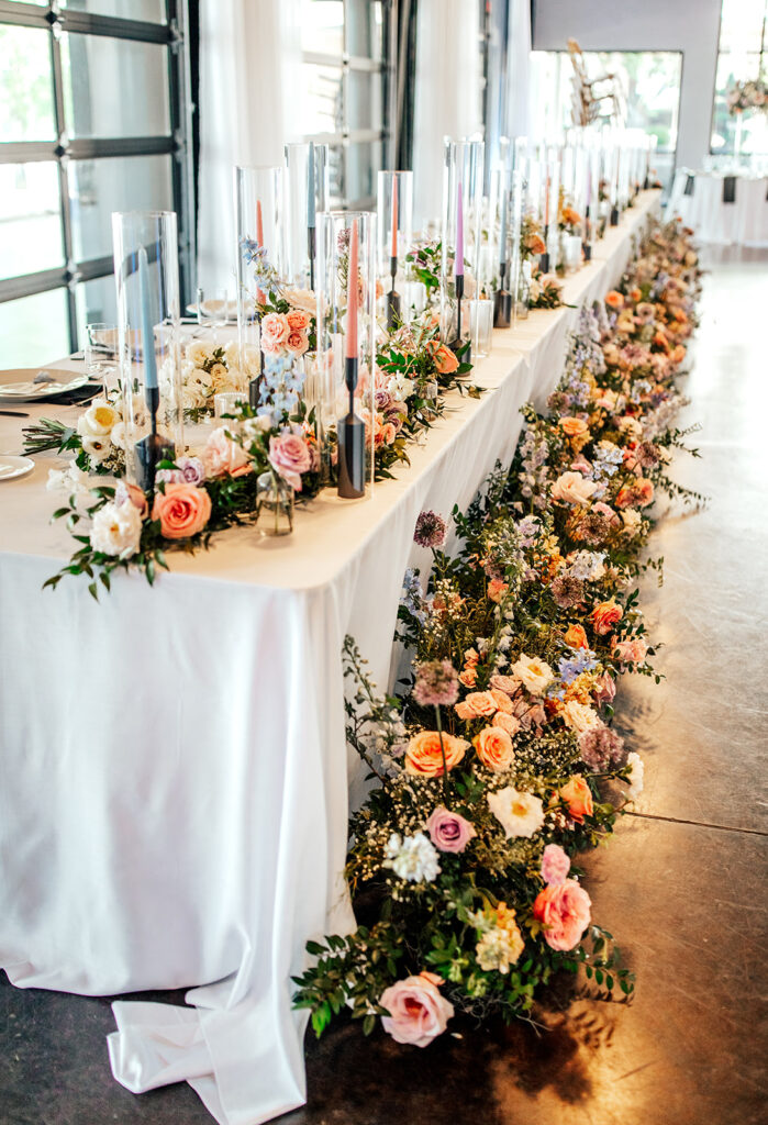 Lush floral head table for wedding reception in Nashville, TN. Colorful and fun wedding flowers for summer wedding reception. Florals running along the base of the head table for wedding reception with table floral garland. Fun colorful wedding candles. Design by Rosemary & Finch in Nashville, TN.