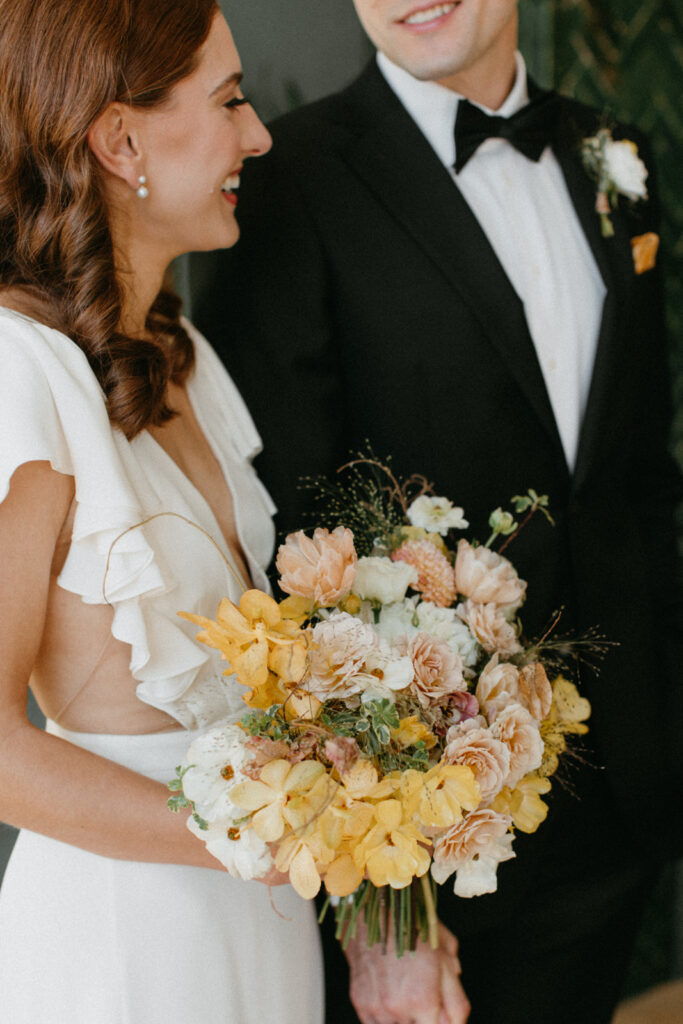 Artful bridal bouquet in yellow tones with cream accents brings unique floral design to fall wedding. Dainty blooms of tulips, spray roses, butterfly ranunculus create colorful fall floral design. Wedding party florals for fall wedding in unique yellow, cream, and toffee colors. Urban fall wedding with modern romantic details. Destination wedding floral designer. Design by Rosemary & Finch Floral Design in Nashville, TN. 
