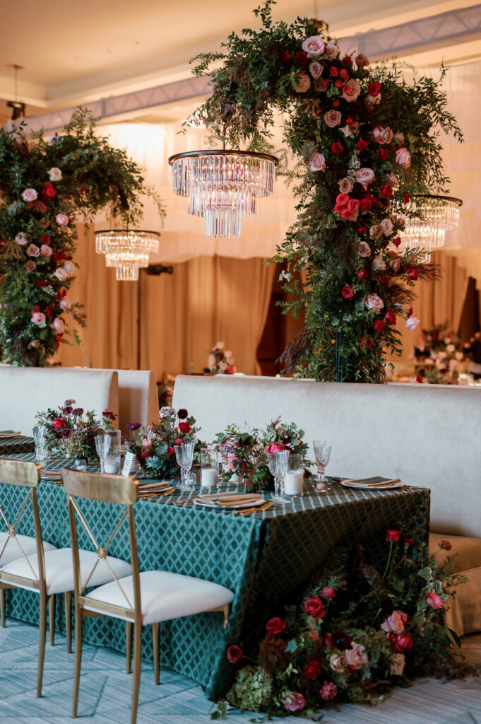 Standing chandeliers covered in lush florals for winter wedding at the Four Seasons. Jewel tone wedding reception florals with reception table floral cascades and table meadows. Winter floral colors in ruby, blush, plum, and magenta. Accent arrangement runners for wedding reception tablescapes. Design by Rosemary & Finch Floral Design.