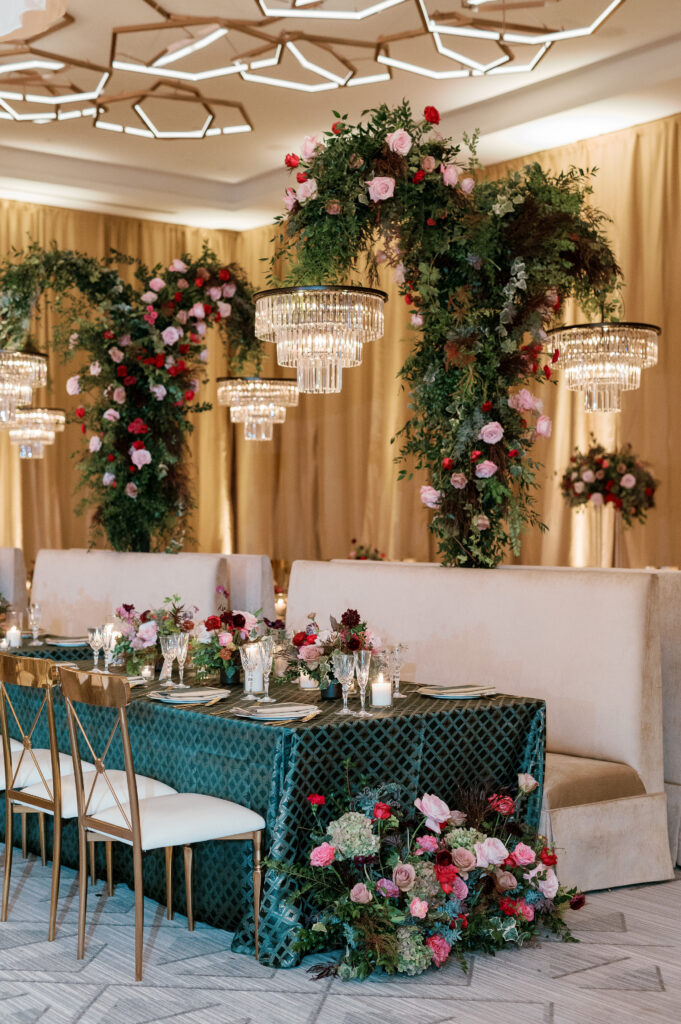 Standing chandeliers covered in lush florals for winter wedding at the Four Seasons. Jewel tone wedding reception florals with reception table floral cascades and table meadows. Winter floral colors in ruby, blush, plum, and magenta. Accent arrangement runners for wedding reception tablescapes. Design by Rosemary & Finch Floral Design.