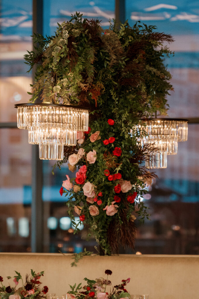 Standing chandeliers covered in lush florals for winter wedding at the Four Seasons. Jewel tone wedding reception florals with reception table floral cascades and table meadows. Winter floral colors in ruby, blush, plum, and magenta. Accent arrangement runners for wedding reception tablescapes. Design by Rosemary & Finch Floral Design.