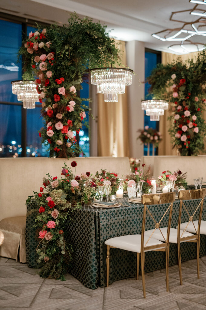 Standing chandeliers covered in lush florals for winter wedding at the Four Seasons. Jewel tone wedding reception florals with reception table floral cascades and table meadows. Winter floral colors in ruby, blush, plum, and magenta. Accent arrangement runners for wedding reception tablescapes. Design by Rosemary & Finch Floral Design.