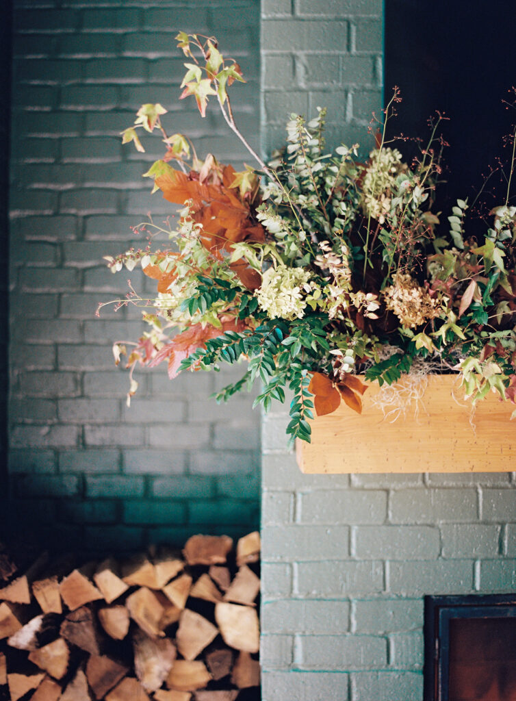 Fireplace fall floral installation for bridesmaids’ luncheon. Outdoor fireplace event décor with fall foliage and green painted brick. Fall event décor with fall foliage and florals in cream, copper, rust, and natural greens. Design by Rosemary & Finch Floral & Finch. 