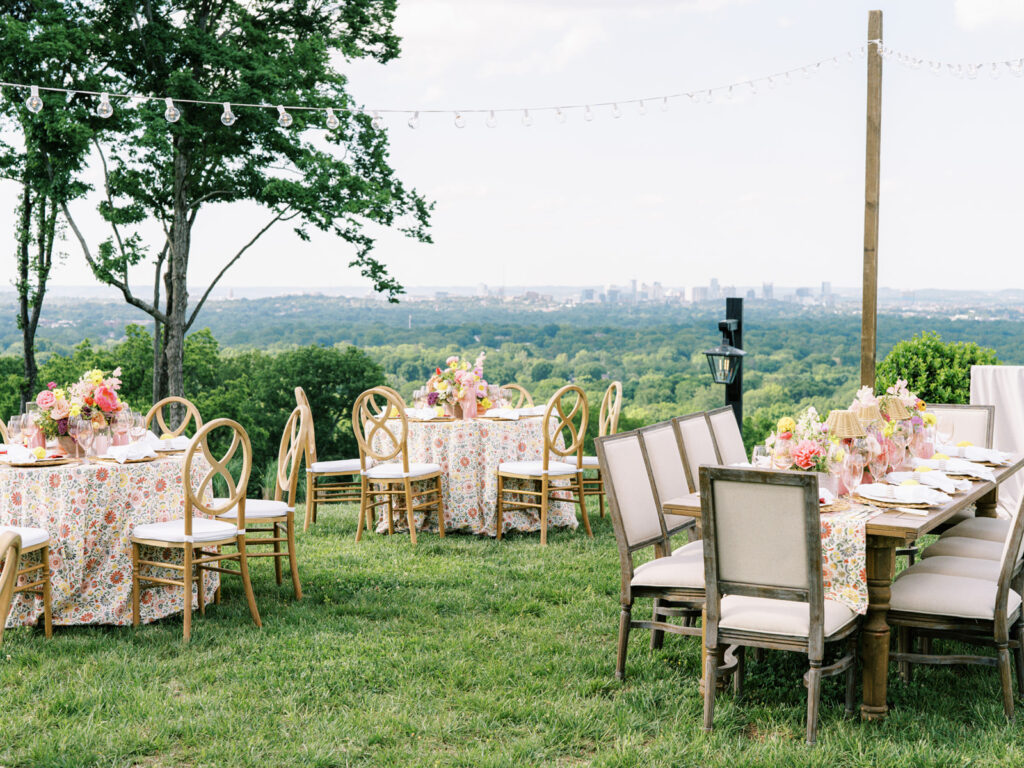 Elegantly set outdoor table with a picturesque view of the downtown Nashville skyline. Tables adorned with floral tablecloths featuring a colorful and intricate pattern. Each table has floral centerpieces with a mix of pink, peach, and yellow flowers, adding a vibrant touch to the setting. Floral design for garden inspired birthday party. Peony inspired floral design with bright spring florals. Design by Rosemary & Finch Floral Design in Nashville, TN. 