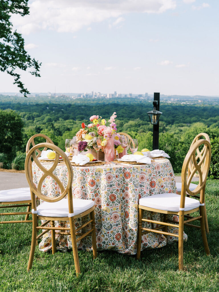 Elegantly set outdoor table with a picturesque view of the downtown Nashville skyline. Tables adorned with floral tablecloths featuring a colorful and intricate pattern. Each table has floral centerpieces with a mix of pink, peach, and yellow flowers, adding a vibrant touch to the setting. Floral design for garden inspired birthday party. Peony inspired floral design with bright spring florals. Design by Rosemary & Finch Floral Design in Nashville, TN. 