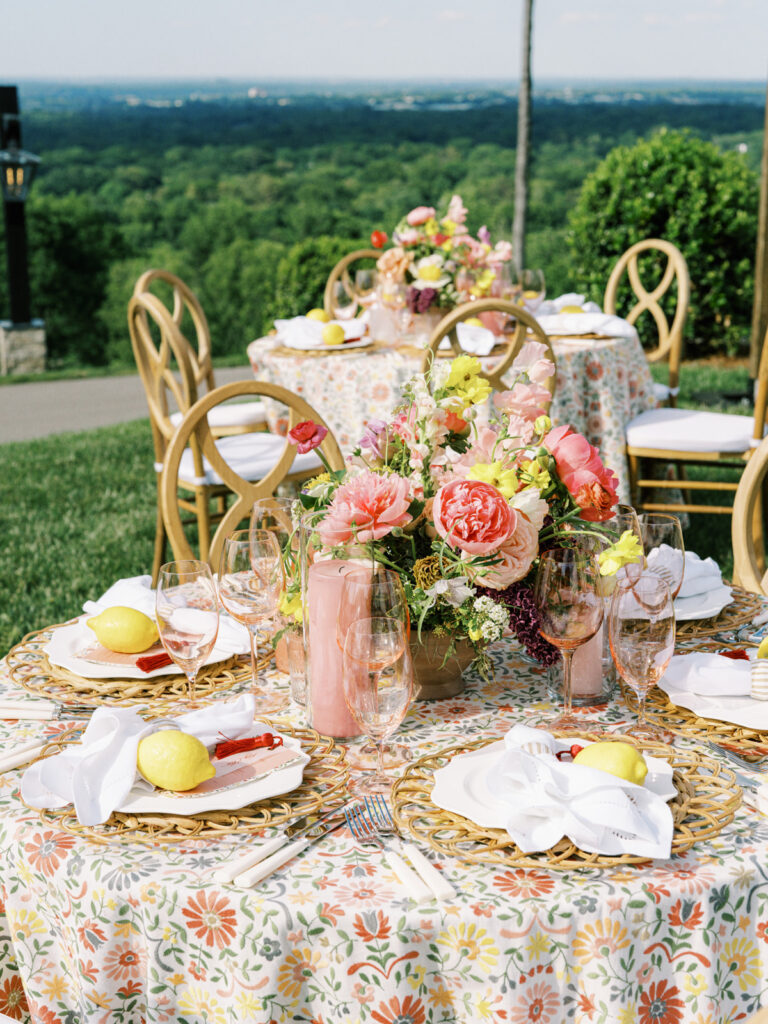 Elegantly set outdoor table with a picturesque view of the downtown Nashville skyline. Tables adorned with floral tablecloths featuring a colorful and intricate pattern. Each table has floral centerpieces with a mix of pink, peach, and yellow flowers, adding a vibrant touch to the setting. Floral design for garden inspired birthday party. Peony inspired floral design with bright spring florals. Design by Rosemary & Finch Floral Design in Nashville, TN. 