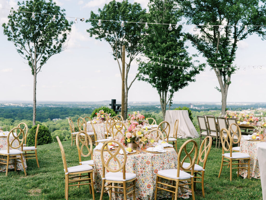 Elegantly set outdoor table with a picturesque view of the downtown Nashville skyline. Tables adorned with floral tablecloths featuring a colorful and intricate pattern. Each table has floral centerpieces with a mix of pink, peach, and yellow flowers, adding a vibrant touch to the setting. Floral design for garden inspired birthday party. Peony inspired floral design with bright spring florals. Design by Rosemary & Finch Floral Design in Nashville, TN. 