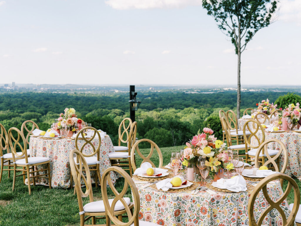 Elegantly set outdoor table with a picturesque view of the downtown Nashville skyline. Tables adorned with floral tablecloths featuring a colorful and intricate pattern. Each table has floral centerpieces with a mix of pink, peach, and yellow flowers, adding a vibrant touch to the setting. Floral design for garden inspired birthday party. Peony inspired floral design with bright spring florals. Design by Rosemary & Finch Floral Design in Nashville, TN. 