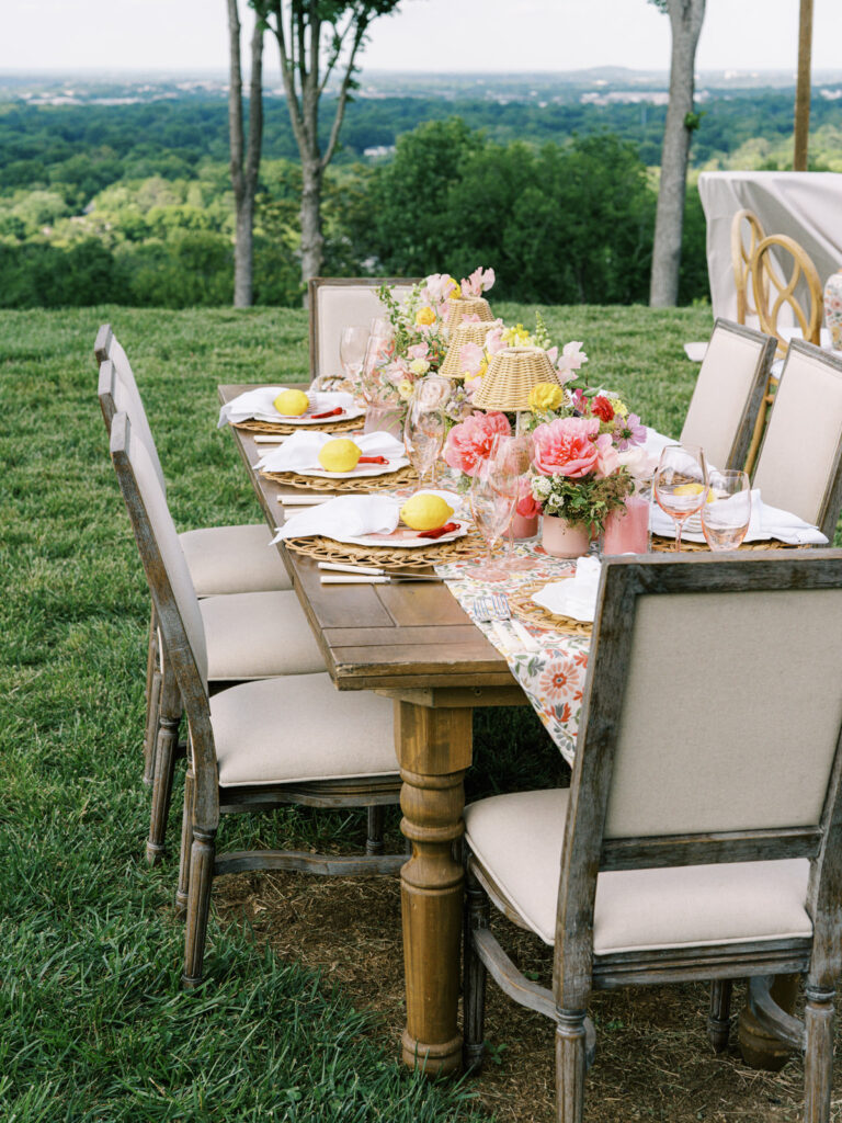 Elegantly set outdoor table with a picturesque view of the downtown Nashville skyline. Tables adorned with floral tablecloths featuring a colorful and intricate pattern. Each table has floral centerpieces with a mix of pink, peach, and yellow flowers, adding a vibrant touch to the setting. Floral design for garden inspired birthday party. Peony inspired floral design with bright spring florals. Design by Rosemary & Finch Floral Design in Nashville, TN. 