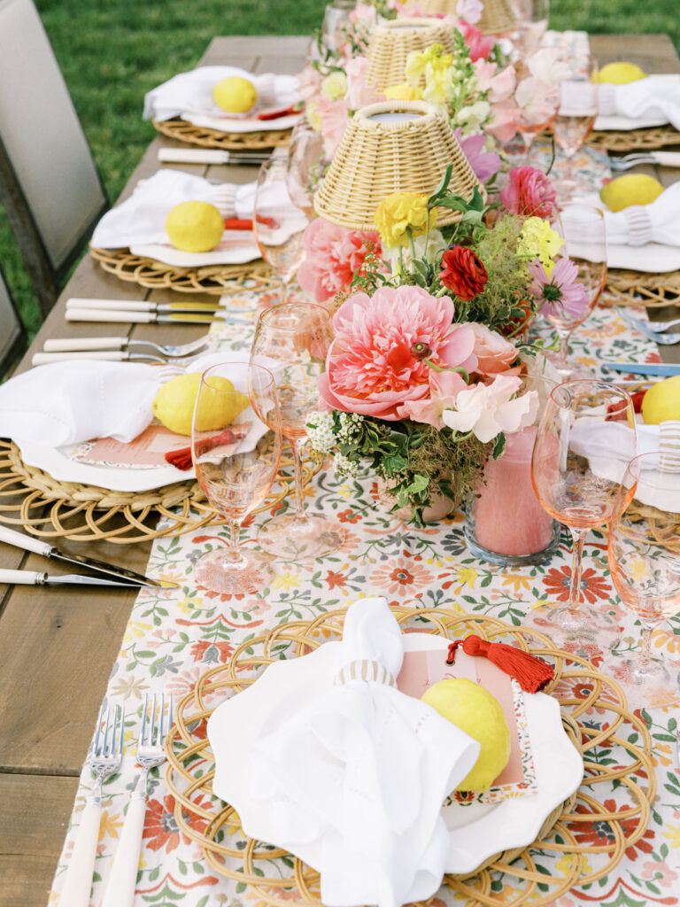 Elegantly set outdoor table with a picturesque view of the downtown Nashville skyline. Tables adorned with floral tablecloths featuring a colorful and intricate pattern. Each table has floral centerpieces with a mix of pink, peach, and yellow flowers, adding a vibrant touch to the setting. Floral design for garden inspired birthday party. Peony inspired floral design with bright spring florals. Design by Rosemary & Finch Floral Design in Nashville, TN. 