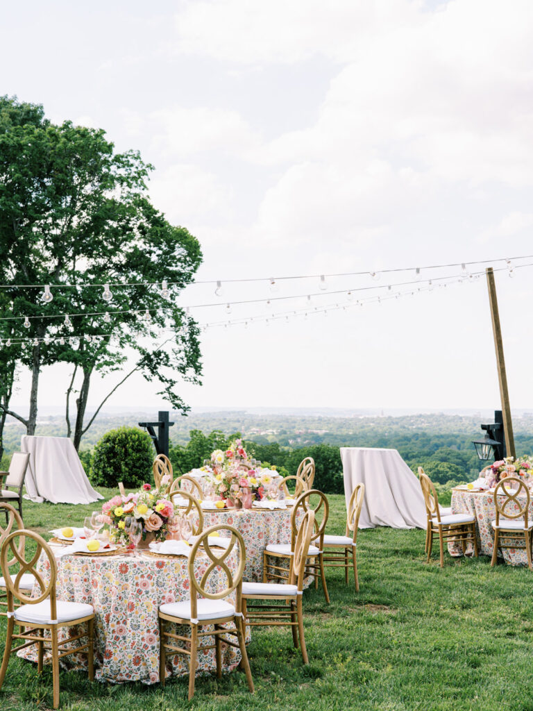 Elegantly set outdoor table with a picturesque view of the downtown Nashville skyline. Tables adorned with floral tablecloths featuring a colorful and intricate pattern. Each table has floral centerpieces with a mix of pink, peach, and yellow flowers, adding a vibrant touch to the setting. Floral design for garden inspired birthday party. Peony inspired floral design with bright spring florals. Design by Rosemary & Finch Floral Design in Nashville, TN. 