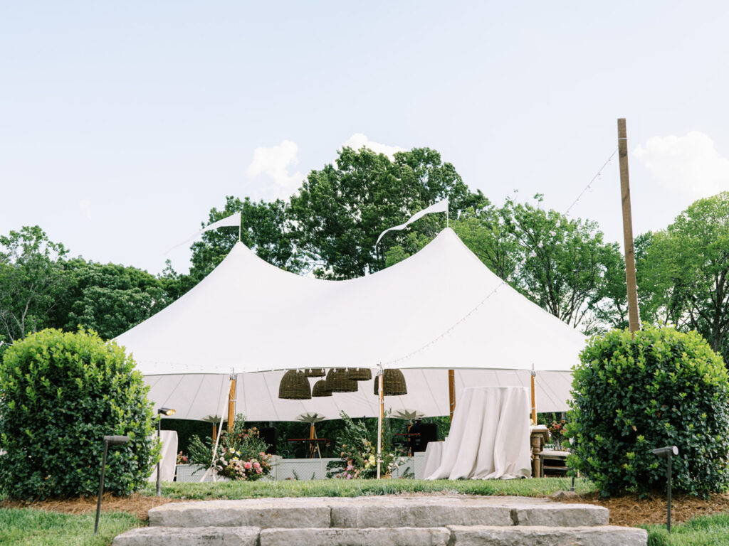 Beautifully set up sailcloth tent for a birthday event in a lush outdoor setting. Tented outdoor event features a peaked design with flowing white fabric that creates an elegant and airy atmosphere. Garden inspired event feature basket pendant lighting and vibrant spring florals. The overall scene exudes a sophisticated and inviting atmosphere, perfect for an elegant outdoor gathering. Design by Rosemary & Finch Floral Design in Nashville, TN.