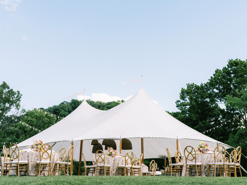 Beautifully set up sailcloth tent for a birthday event in a lush outdoor setting. Tented outdoor event features a peaked design with flowing white fabric that creates an elegant and airy atmosphere. Garden inspired event feature basket pendant lighting and vibrant spring florals. The overall scene exudes a sophisticated and inviting atmosphere, perfect for an elegant outdoor gathering. Design by Rosemary & Finch Floral Design in Nashville, TN.