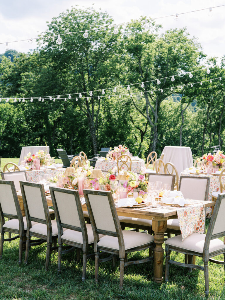 Elegantly set outdoor table with a picturesque view of the downtown Nashville skyline. Tables adorned with floral tablecloths featuring a colorful and intricate pattern. Each table has floral centerpieces with a mix of pink, peach, and yellow flowers, adding a vibrant touch to the setting. Floral design for garden inspired birthday party. Peony inspired floral design with bright spring florals. Design by Rosemary & Finch Floral Design in Nashville, TN. 