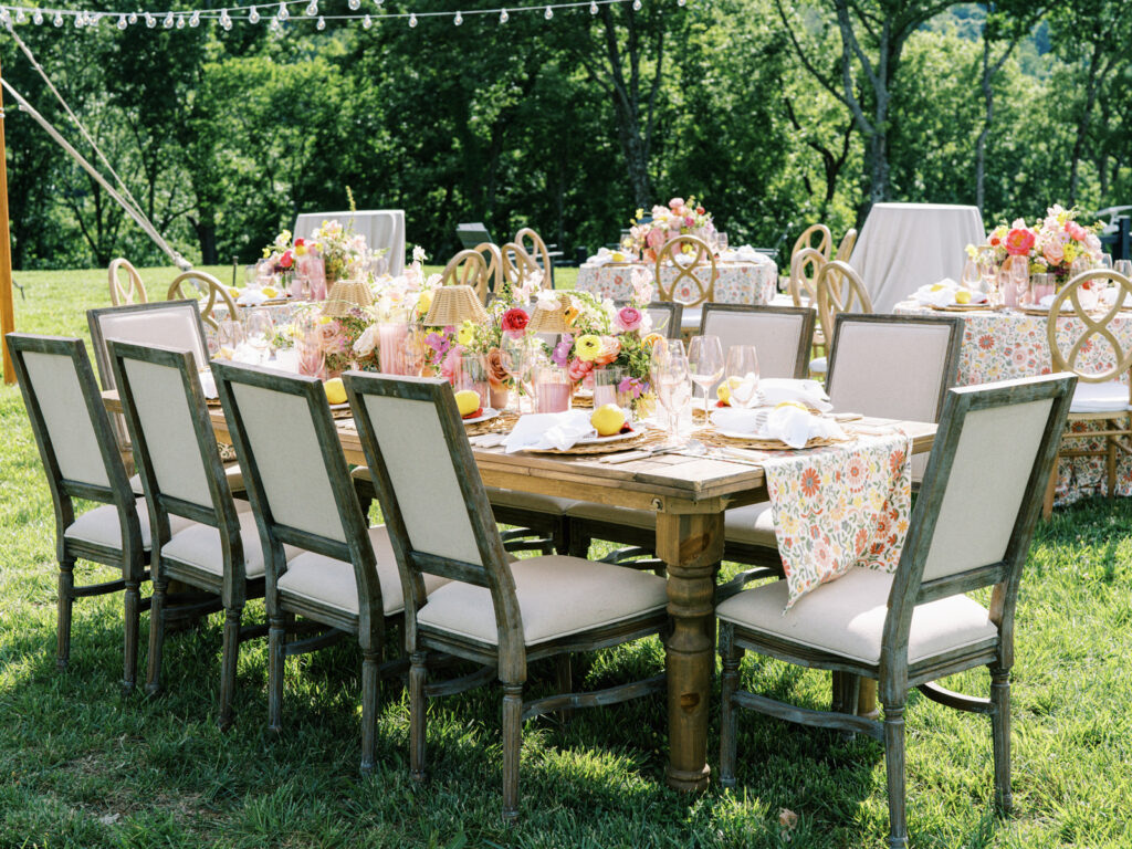 Elegantly set outdoor table with a picturesque view of the downtown Nashville skyline. Tables adorned with floral tablecloths featuring a colorful and intricate pattern. Each table has floral centerpieces with a mix of pink, peach, and yellow flowers, adding a vibrant touch to the setting. Floral design for garden inspired birthday party. Peony inspired floral design with bright spring florals. Design by Rosemary & Finch Floral Design in Nashville, TN. 