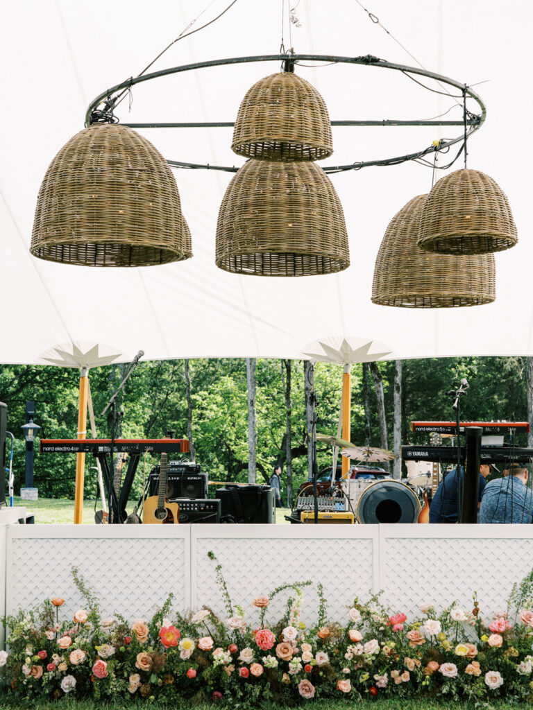 Flowers arranged to create a lush, vibrant border that complements the white lattice stage facade. The selection of flowers, ranging from roses to peonies and other seasonal blooms, adds texture and depth. Installation features a mix of heights to create a natural, garden-like feel. Sailcloth private evet with basket pendant lighting and spring florals. Design by Rosemary & Finch Floral Design. 