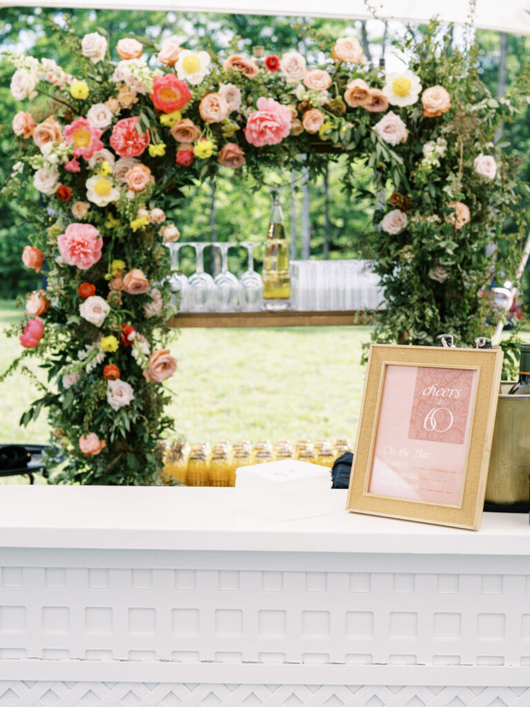 White lattice bar under sailcloth tent for private birthday party event. Beautiful floral installation forms an archway, featuring a variety of flowers in shades of pink, peach, yellow, and white. Framed menus along with additional floral accents tie in with the main floral arch behind bar. Florals serve as a beautiful backdrop and adds a festive and cheerful vibe to the atmosphere. Design by Rosemary & Finch Floral Design in Nashville, TN. 