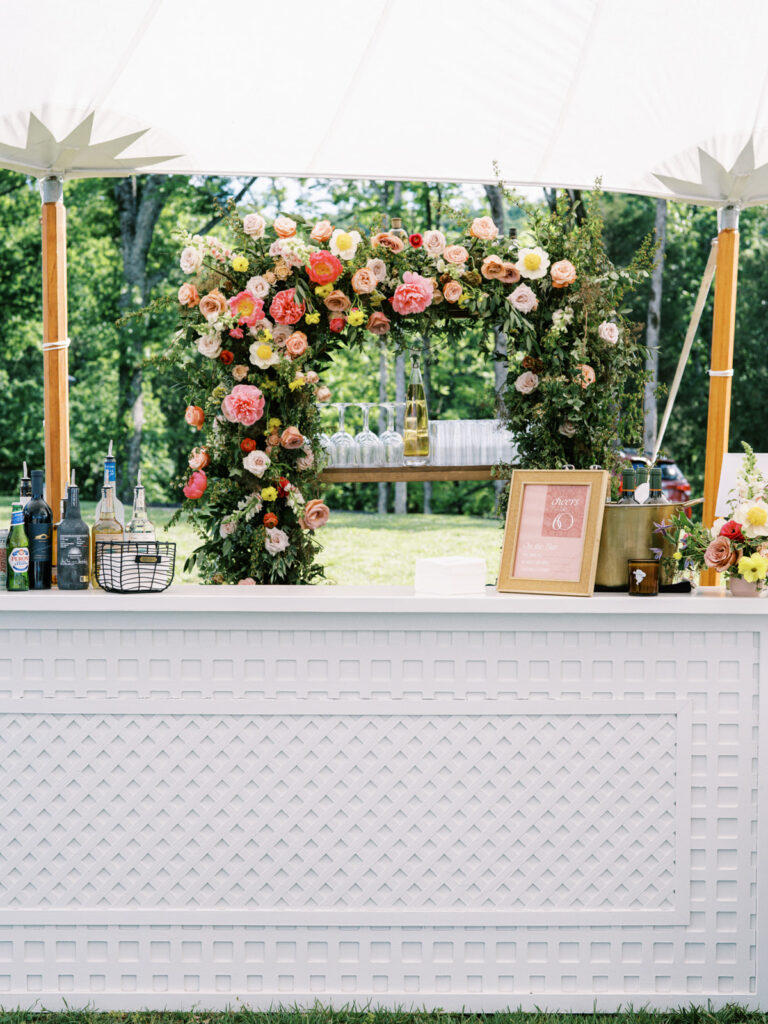 White lattice bar under sailcloth tent for private birthday party event. Beautiful floral installation forms an archway, featuring a variety of flowers in shades of pink, peach, yellow, and white. Framed menus along with additional floral accents tie in with the main floral arch behind bar. Florals serve as a beautiful backdrop and adds a festive and cheerful vibe to the atmosphere. Design by Rosemary & Finch Floral Design in Nashville, TN. 