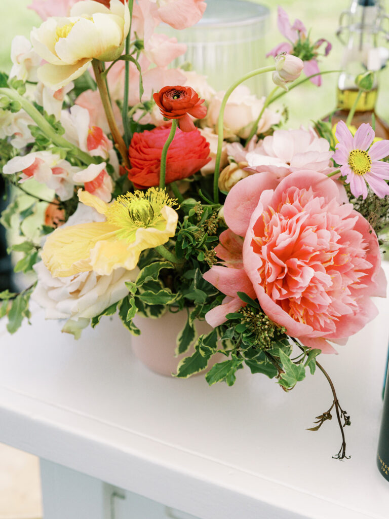 Outdoor garden-inspired event for birthday party. Medium floral arrangements adorn the coffee table and interactive tables of event. The arrangement consists of a variety of colorful flowers, including pink peonies, yellow and peach roses, and other delicate blooms. The combination of modern furniture, vibrant florals, and comfortable seating are perfect for a memorable outdoor event. Design by Rosemary & Finch Floral Design in Nashville, TN.