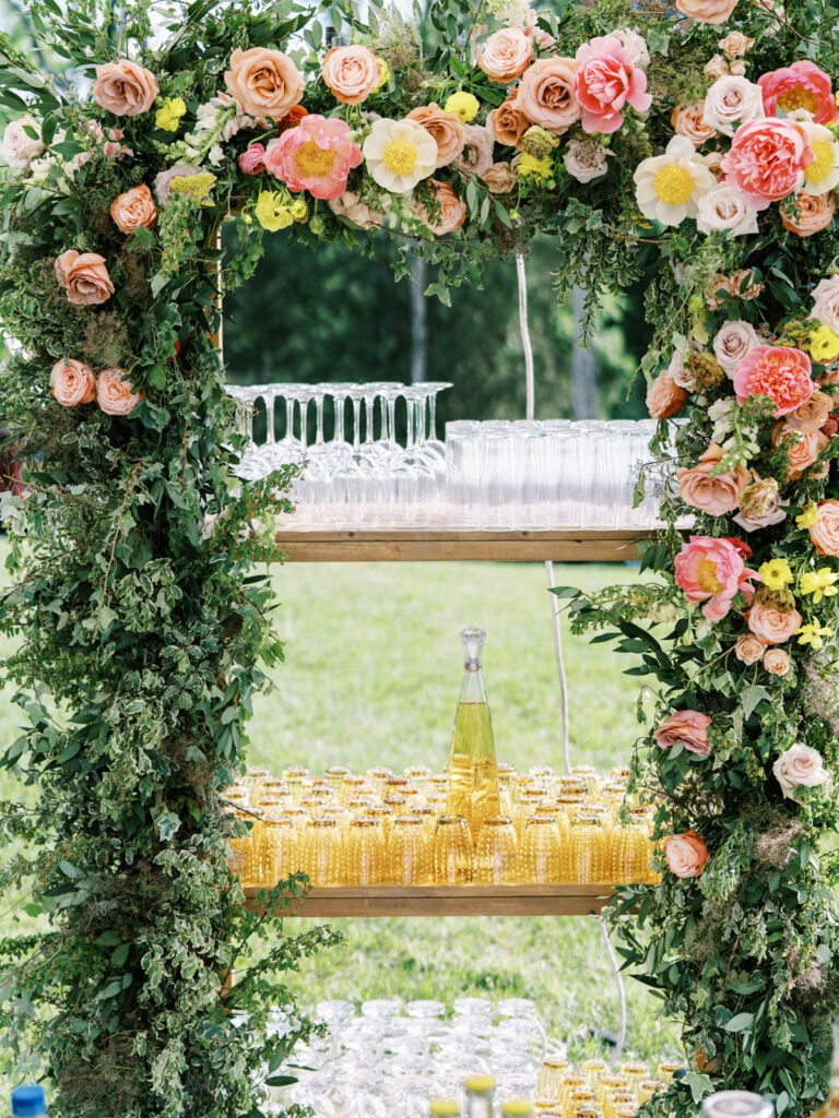 White lattice bar under sailcloth tent for private birthday party event. Beautiful floral installation forms an archway, featuring a variety of flowers in shades of pink, peach, yellow, and white. Framed menus along with additional floral accents tie in with the main floral arch behind bar. Florals serve as a beautiful backdrop and adds a festive and cheerful vibe to the atmosphere. Design by Rosemary & Finch Floral Design in Nashville, TN. 