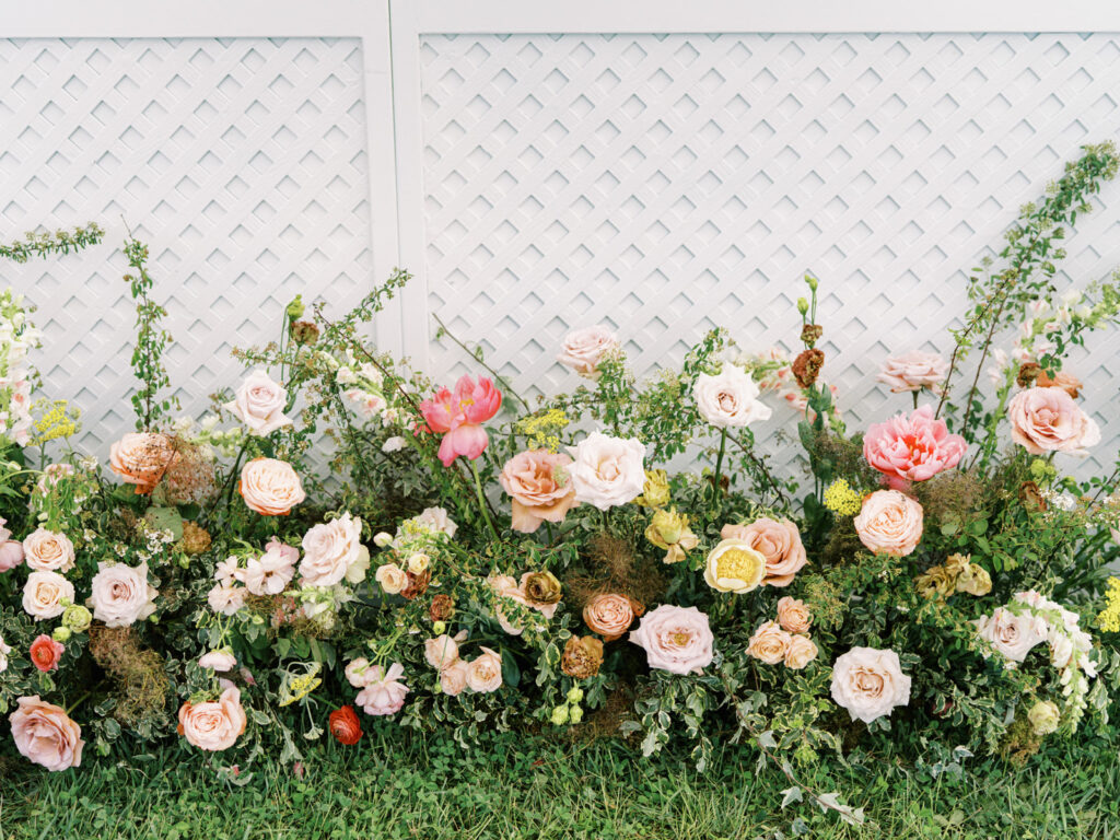 Flowers arranged to create a lush, vibrant border that complements the white lattice stage facade. The selection of flowers, ranging from roses to peonies and other seasonal blooms, adds texture and depth. Installation features a mix of heights to create a natural, garden-like feel. Sailcloth private evet with basket pendant lighting and spring florals. Design by Rosemary & Finch Floral Design. 