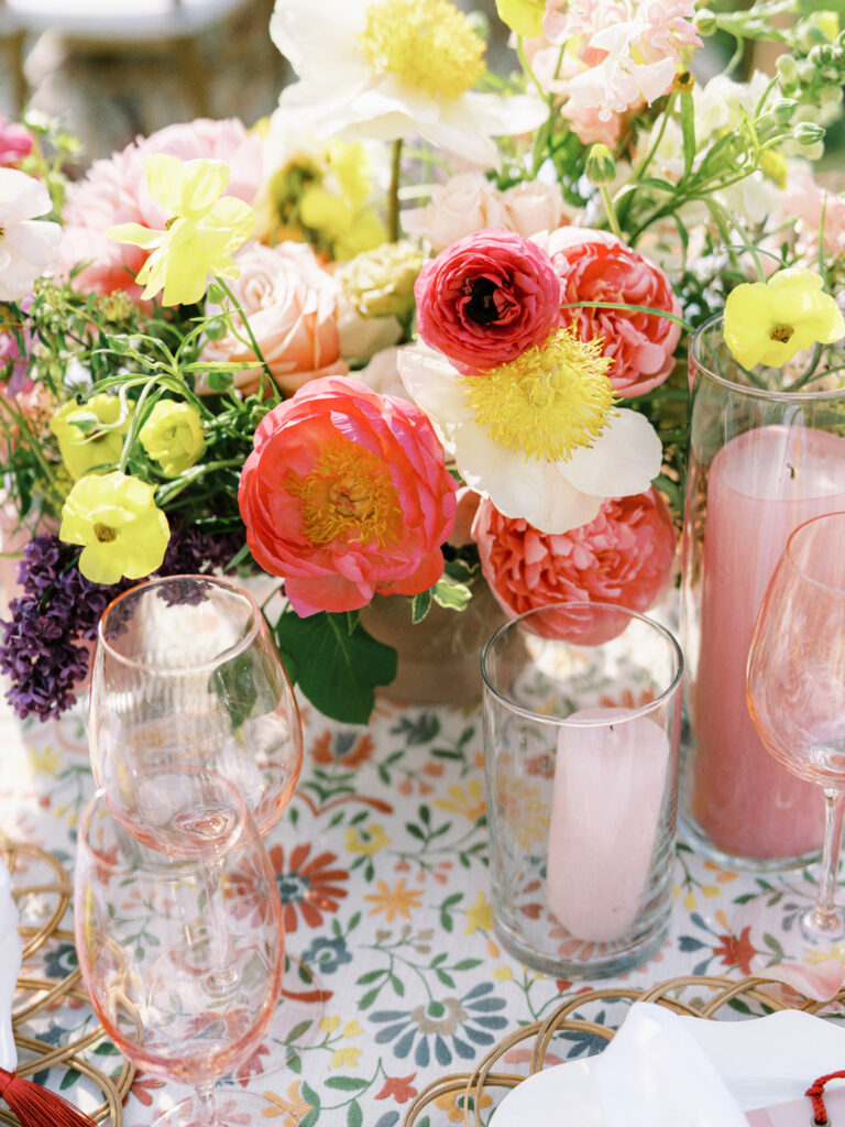 Elegantly set outdoor table with a picturesque view of the downtown Nashville skyline. Tables adorned with floral tablecloths featuring a colorful and intricate pattern. Each table has floral centerpieces with a mix of pink, peach, and yellow flowers, adding a vibrant touch to the setting. Floral design for garden inspired birthday party. Peony inspired floral design with bright spring florals. Design by Rosemary & Finch Floral Design in Nashville, TN. 