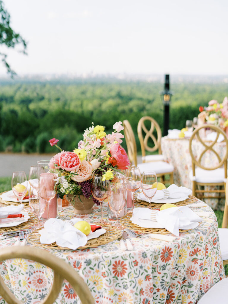 Elegantly set outdoor table with a picturesque view of the downtown Nashville skyline. Tables adorned with floral tablecloths featuring a colorful and intricate pattern. Each table has floral centerpieces with a mix of pink, peach, and yellow flowers, adding a vibrant touch to the setting. Floral design for garden inspired birthday party. Peony inspired floral design with bright spring florals. Design by Rosemary & Finch Floral Design in Nashville, TN. 