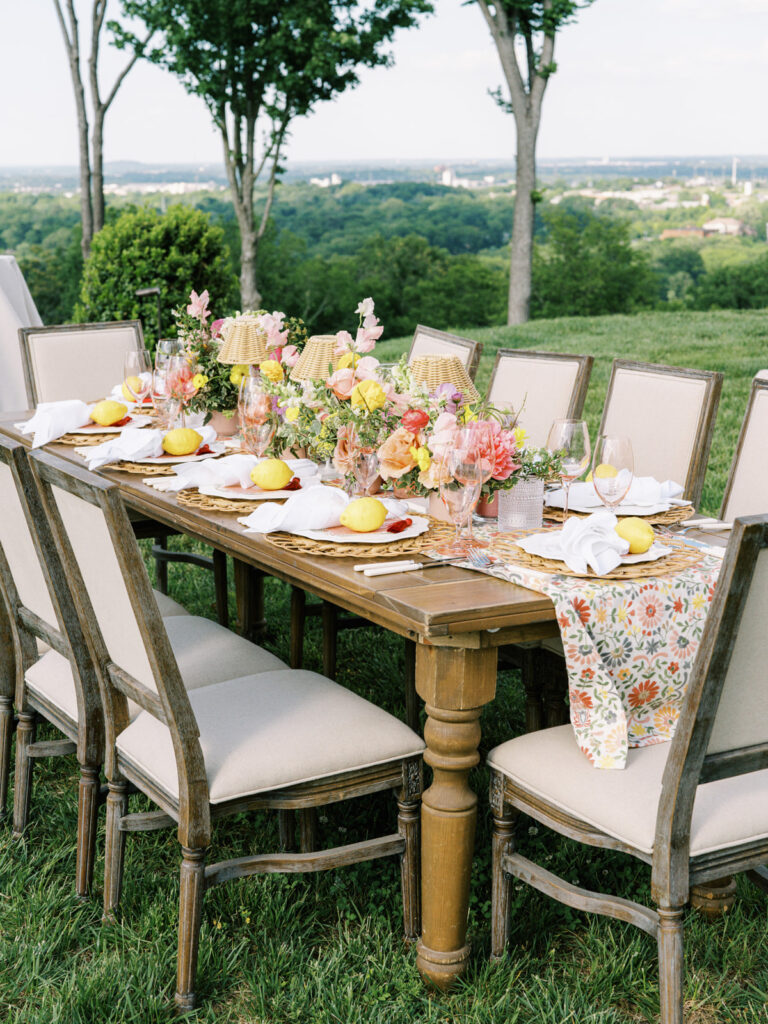 Elegantly set outdoor table with a picturesque view of the downtown Nashville skyline. Tables adorned with floral tablecloths featuring a colorful and intricate pattern. Each table has floral centerpieces with a mix of pink, peach, and yellow flowers, adding a vibrant touch to the setting. Floral design for garden inspired birthday party. Peony inspired floral design with bright spring florals. Design by Rosemary & Finch Floral Design in Nashville, TN. 