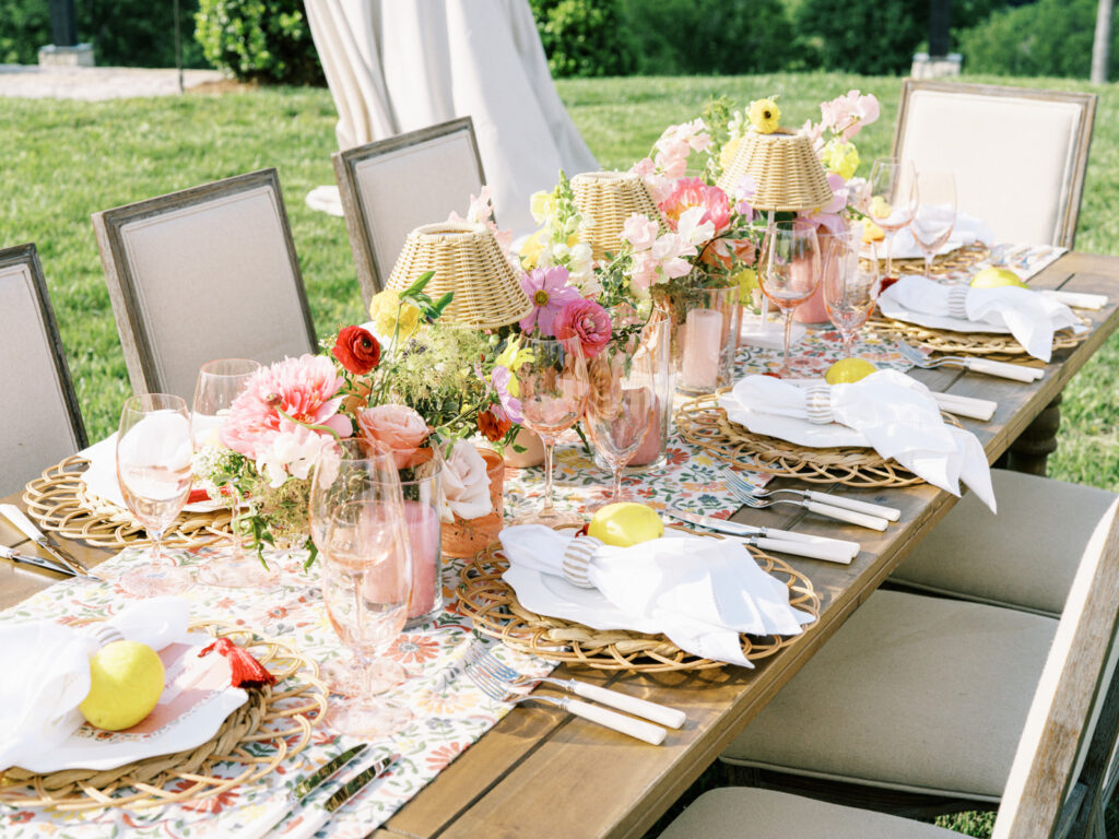 Elegantly set outdoor table with a picturesque view of the downtown Nashville skyline. Tables adorned with floral tablecloths featuring a colorful and intricate pattern. Each table has floral centerpieces with a mix of pink, peach, and yellow flowers, adding a vibrant touch to the setting. Floral design for garden inspired birthday party. Peony inspired floral design with bright spring florals. Design by Rosemary & Finch Floral Design in Nashville, TN. 