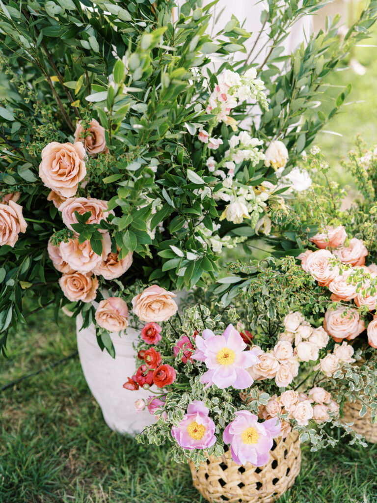 The floral display features a variety of blooms in soft, romantic hues, including peach roses, delicate white flowers, and vibrant red and pink blossoms. Moab roses and peonies are abundant and full, adding a lush and luxurious feel to the arrangement. Single product basket and urns allow the natural beauty of the flowers to stand out. Fresh, garden-like atmosphere created at the entrance of the sailcloth tent. Design by Rosemary & Finch Floral Design.