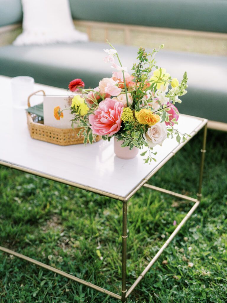 Outdoor garden-inspired event for birthday party. Medium floral arrangements adorn the coffee table and interactive tables of event. The arrangement consists of a variety of colorful flowers, including pink peonies, yellow and peach roses, and other delicate blooms. The combination of modern furniture, vibrant florals, and comfortable seating are perfect for a memorable outdoor event. Design by Rosemary & Finch Floral Design in Nashville, TN.