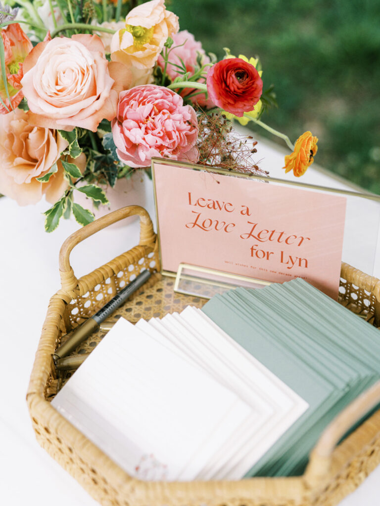 Outdoor garden-inspired event for birthday party. Medium floral arrangements adorn the coffee table and interactive tables of event. The arrangement consists of a variety of colorful flowers, including pink peonies, yellow and peach roses, and other delicate blooms. The combination of modern furniture, vibrant florals, and comfortable seating are perfect for a memorable outdoor event. Design by Rosemary & Finch Floral Design in Nashville, TN.