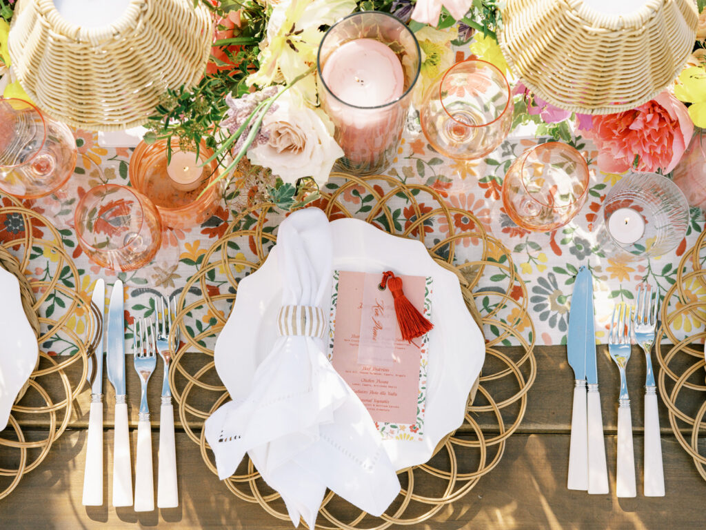 Elegantly set outdoor table with a picturesque view of the downtown Nashville skyline. Tables adorned with floral tablecloths featuring a colorful and intricate pattern. Each table has floral centerpieces with a mix of pink, peach, and yellow flowers, adding a vibrant touch to the setting. Floral design for garden inspired birthday party. Peony inspired floral design with bright spring florals. Design by Rosemary & Finch Floral Design in Nashville, TN. 