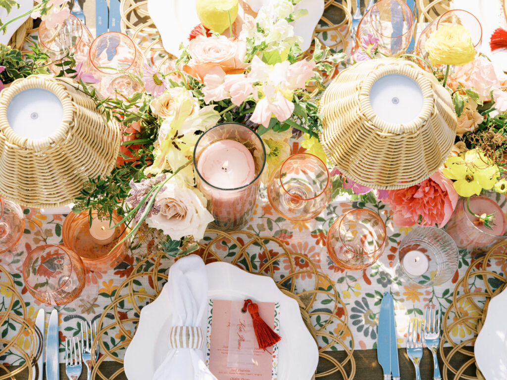 Elegantly set outdoor table with a picturesque view of the downtown Nashville skyline. Tables adorned with floral tablecloths featuring a colorful and intricate pattern. Each table has floral centerpieces with a mix of pink, peach, and yellow flowers, adding a vibrant touch to the setting. Floral design for garden inspired birthday party. Peony inspired floral design with bright spring florals. Design by Rosemary & Finch Floral Design in Nashville, TN. 