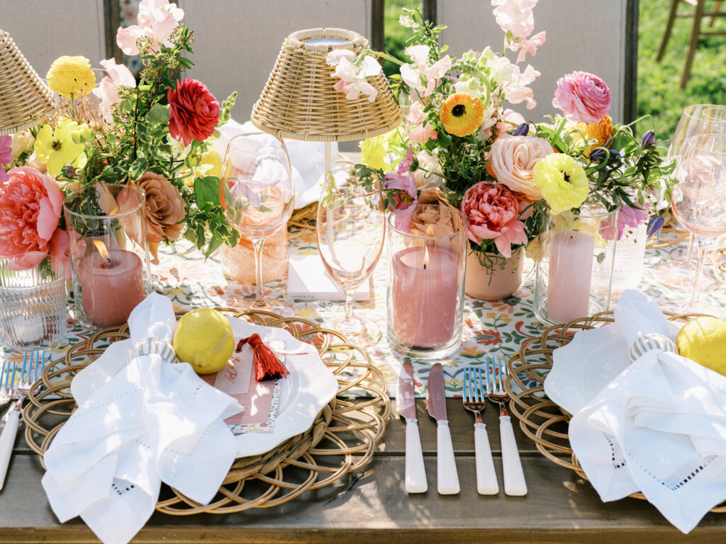 Elegantly set outdoor table with a picturesque view of the downtown Nashville skyline. Tables adorned with floral tablecloths featuring a colorful and intricate pattern. Each table has floral centerpieces with a mix of pink, peach, and yellow flowers, adding a vibrant touch to the setting. Floral design for garden inspired birthday party. Peony inspired floral design with bright spring florals. Design by Rosemary & Finch Floral Design in Nashville, TN. 