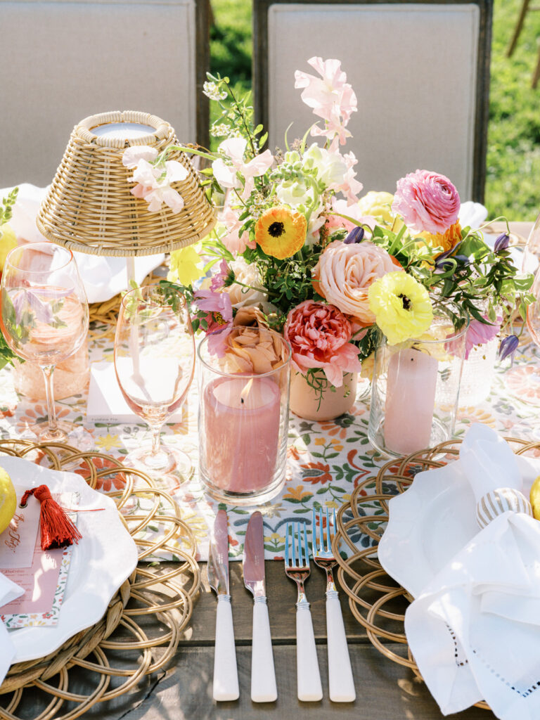 Elegantly set outdoor table with a picturesque view of the downtown Nashville skyline. Tables adorned with floral tablecloths featuring a colorful and intricate pattern. Each table has floral centerpieces with a mix of pink, peach, and yellow flowers, adding a vibrant touch to the setting. Floral design for garden inspired birthday party. Peony inspired floral design with bright spring florals. Design by Rosemary & Finch Floral Design in Nashville, TN. 