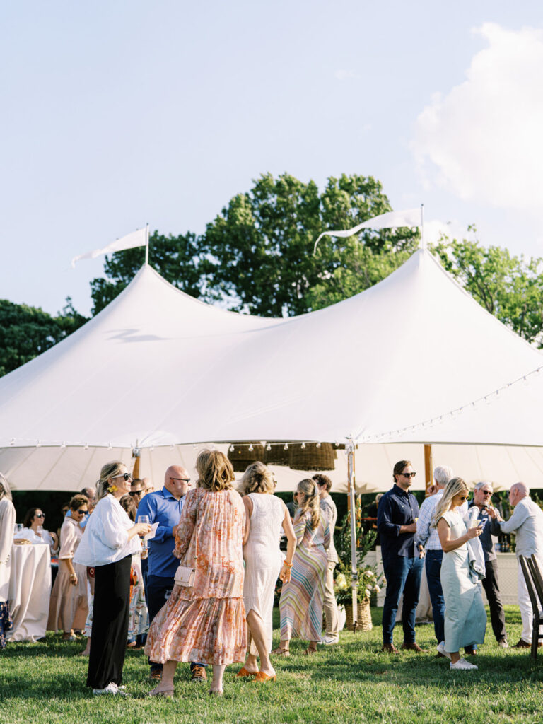 Beautifully set up sailcloth tent for a birthday event in a lush outdoor setting. Tented outdoor event features a peaked design with flowing white fabric that creates an elegant and airy atmosphere. Garden inspired event feature basket pendant lighting and vibrant spring florals. The overall scene exudes a sophisticated and inviting atmosphere, perfect for an elegant outdoor gathering. Design by Rosemary & Finch Floral Design in Nashville, TN.