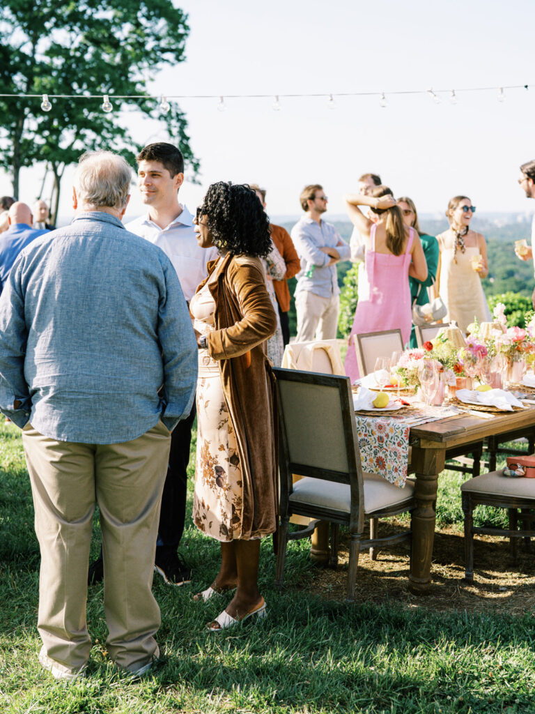 Elegantly set outdoor table with a picturesque view of the downtown Nashville skyline. Tables adorned with floral tablecloths featuring a colorful and intricate pattern. Each table has floral centerpieces with a mix of pink, peach, and yellow flowers, adding a vibrant touch to the setting. Floral design for garden inspired birthday party. Peony inspired floral design with bright spring florals. Design by Rosemary & Finch Floral Design in Nashville, TN. 