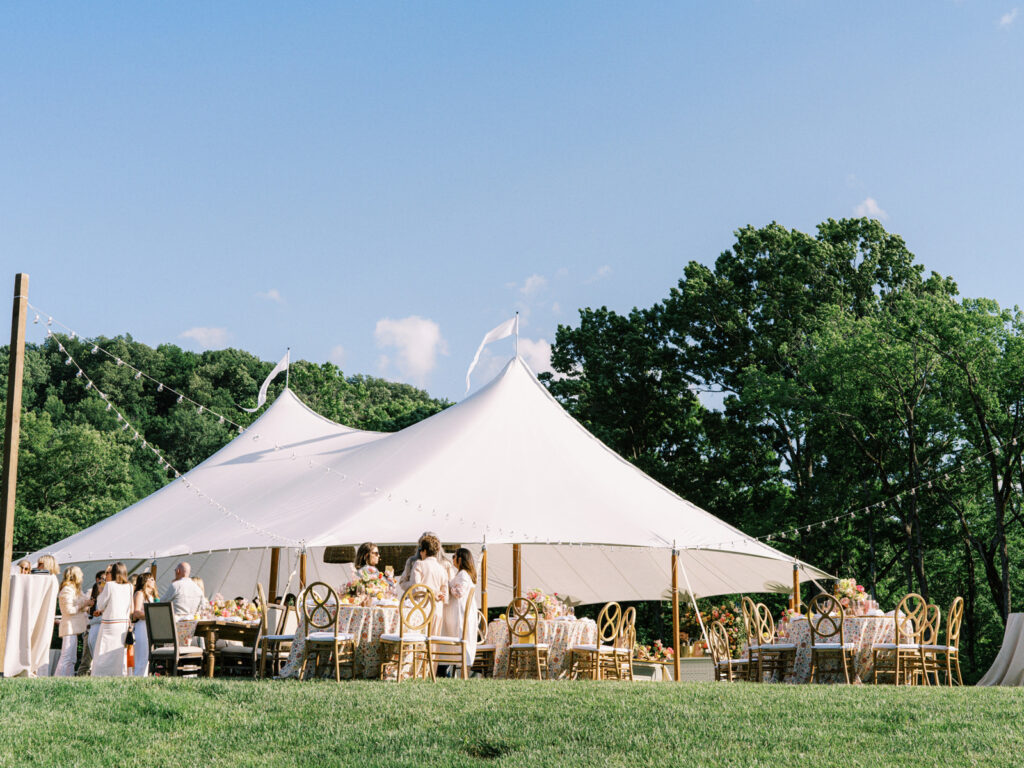 Beautifully set up sailcloth tent for a birthday event in a lush outdoor setting. Tented outdoor event features a peaked design with flowing white fabric that creates an elegant and airy atmosphere. Garden inspired event feature basket pendant lighting and vibrant spring florals. The overall scene exudes a sophisticated and inviting atmosphere, perfect for an elegant outdoor gathering. Design by Rosemary & Finch Floral Design in Nashville, TN.