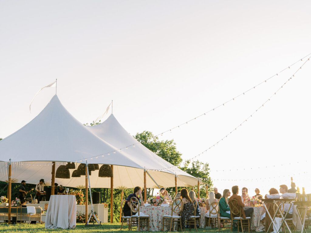 Beautifully set up sailcloth tent for a birthday event in a lush outdoor setting. Tented outdoor event features a peaked design with flowing white fabric that creates an elegant and airy atmosphere. Garden inspired event feature basket pendant lighting and vibrant spring florals. The overall scene exudes a sophisticated and inviting atmosphere, perfect for an elegant outdoor gathering. Design by Rosemary & Finch Floral Design in Nashville, TN.